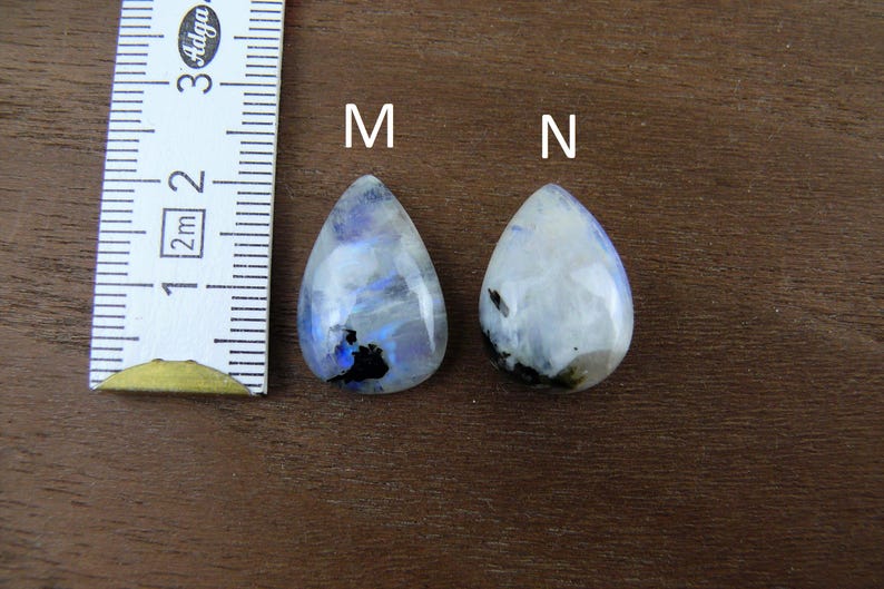 Puede incluir: Dos cabujones de piedra lunar en forma de l&aacute;grima, etiquetados M y N, con una regla para la escala. Las piedras son principalmente blancas con inclusiones azules y negras. La regla muestra medidas en cent&iacute;metros.