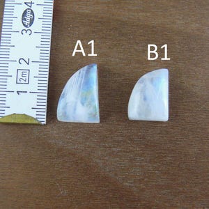 May include: Two white moonstone cabochons with blue iridescence, labeled A1 and B1, positioned next to a measuring tape showing a length of approximately 1.5 inches for the larger stone and 1.25 inches for the smaller stone.