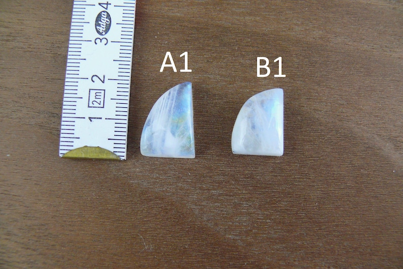 May include: Two white moonstone cabochons with blue iridescence, labeled A1 and B1, positioned next to a measuring tape showing a length of approximately 1.5 inches for the larger stone and 1.25 inches for the smaller stone.