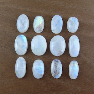 Può includere: Una collezione di dodici cabochon ovali di pietra di luna. Le pietre presentano un colore di base bianco latte con riflessi iridescenti blu e gialli. Le pietre lucide sono disposte su una superficie di legno marrone.