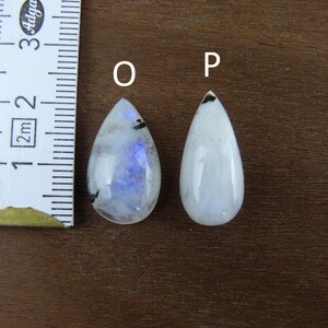 Puede incluir: Dos cabujones de piedra lunar en forma de l&aacute;grima, uno con iridiscencia azul, el otro blanco. Una regla muestra que las piedras miden aproximadamente 2,5 cm de largo. Las piedras est&aacute;n sobre una superficie de madera oscura.