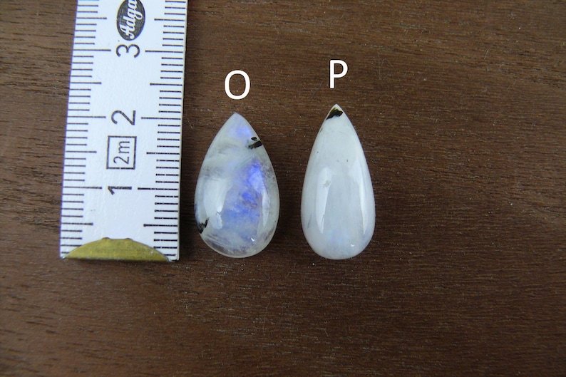 Puede incluir: Dos cabujones de piedra lunar en forma de l&aacute;grima, uno con iridiscencia azul, el otro blanco. Una regla muestra que las piedras miden aproximadamente 2,5 cm de largo. Las piedras est&aacute;n sobre una superficie de madera oscura.