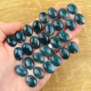 Könnte beinhalten: Eine Sammlung zahlreicher ovaler, polierter Moosachat-Edelsteine. Jeder Stein weist eine tiefdunkelgrüne Farbe mit komplexen, dunklen, moosartigen Einschlüssen auf der glatten, glänzenden Oberfläche der im Cabochon-Schliff gefertigten Steine auf.