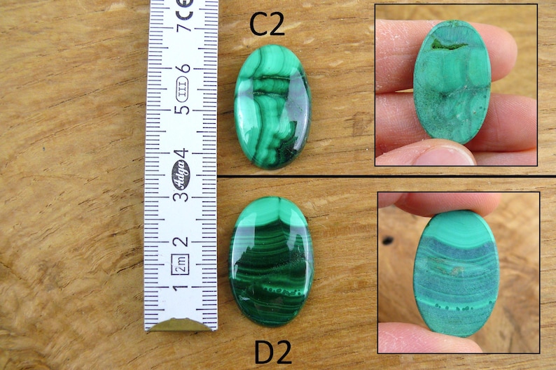 May include: Oval-shaped malachite gemstones with dark green and light green patterns. The image includes a ruler for size reference. The gemstones are displayed on a wooden surface. The text "C2" and "D2" are visible.