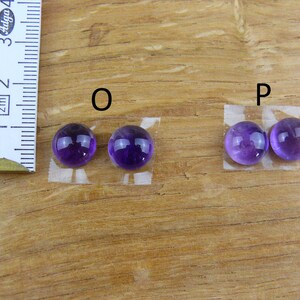 May include: Two pairs of polished, round, purple amethyst gemstones, labeled "O" and "P", are displayed on a wooden surface. A ruler is positioned to the left, indicating measurements. The gemstones are affixed to small pieces of tape.