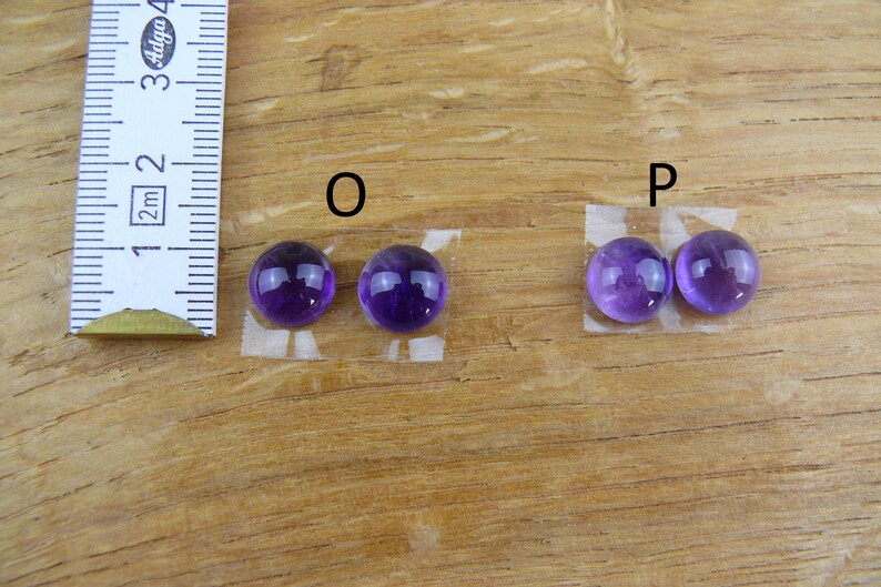 May include: Two pairs of polished, round, purple amethyst gemstones, labeled "O" and "P", are displayed on a wooden surface. A ruler is positioned to the left, indicating measurements. The gemstones are affixed to small pieces of tape.