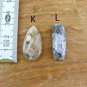 May include: Two polished stone beads labeled K and L next to a ruler showing measurements in centimeters. Bead K is teardrop-shaped with cream, tan, and grey tones. Bead L is a cylindrical shape with mottled grey, white, and dark green patterns.