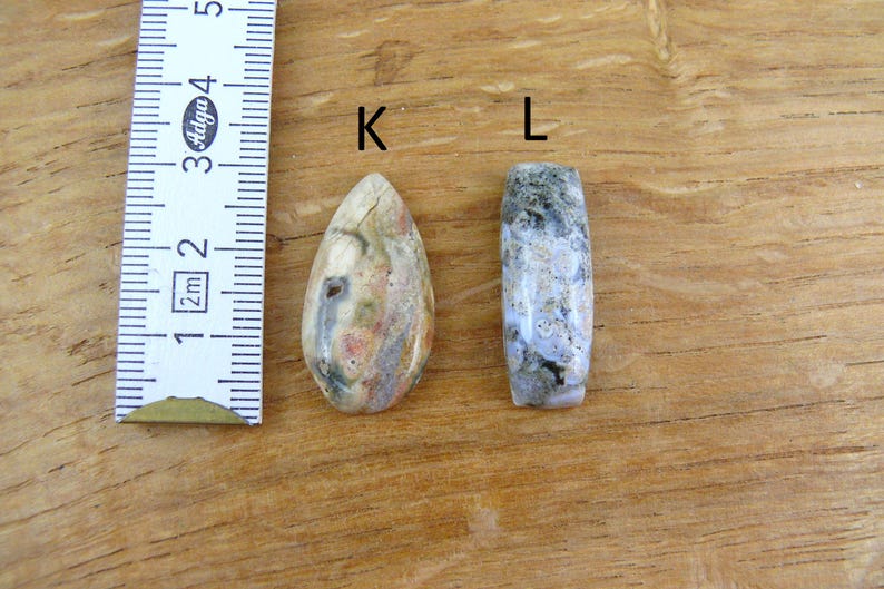 May include: Two polished stone beads labeled K and L next to a ruler showing measurements in centimeters. Bead K is teardrop-shaped with cream, tan, and grey tones. Bead L is a cylindrical shape with mottled grey, white, and dark green patterns.
