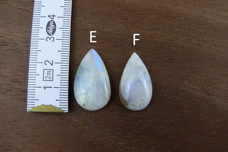 Puede incluir: Dos cabujones de piedra lunar en forma de l&aacute;grima con tonos iridiscentes azules y blancos. Las piedras preciosas se exhiben sobre una superficie de madera junto a una regla. Las piedras lunares miden aproximadamente 2,5 cm de largo.