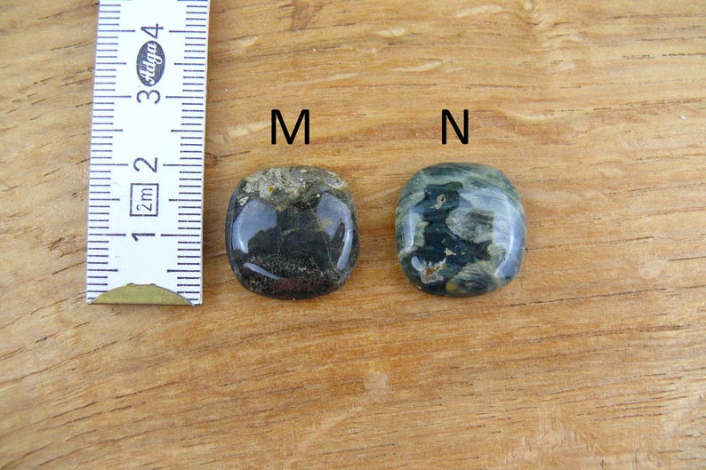 May include: Two polished, rounded stones labeled M and N, positioned next to a white ruler. Stone M is dark gray and black with a textured top edge. Stone N features a mottled pattern of dark green, black, and white. The ruler shows a scale in cm.