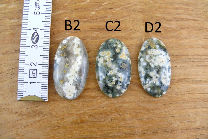 Op de afbeelding: Drie ovaalvormige oceaanjaspis cabochons gelabeld B2, C2 en D2, met witte, grijze en groene cirkelvormige patronen. Een wit meetlint is aan de linkerkant geplaatst en toont een schaal in centimeters voor referentie van de grootte.