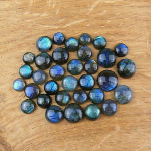Può includere: Una collezione di cabochon di pietre preziose di labradorite lucidata di varie dimensioni. Le pietre mostrano lampi iridescenti di blu, verde e oro su uno sfondo scuro. I cabochon sono disposti su una superficie di legno.
