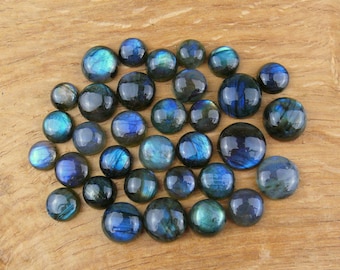 May include: A collection of polished labradorite gemstone cabochons in various sizes. The stones display iridescent flashes of blue, green, and gold against a dark background. The cabochons are arranged on a wooden surface.