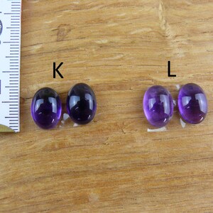 May include: Four oval-shaped, polished amethyst gemstones, two pairs labeled "K" and "L", are displayed on a wooden surface. A ruler in centimeters is positioned to the left, indicating size. The gemstones exhibit a deep purple hue.