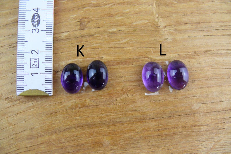 May include: Four oval-shaped, polished amethyst gemstones, two pairs labeled "K" and "L", are displayed on a wooden surface. A ruler in centimeters is positioned to the left, indicating size. The gemstones exhibit a deep purple hue.