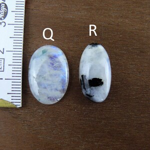 Puede incluir: Dos piedras preciosas de forma ovalada, una con tonos azules y blancos, la otra con inclusiones negras. Se coloca una regla junto a las piedras para comparar el tama&ntilde;o. Las piedras preciosas est&aacute;n sobre una superficie marr&oacute;n.