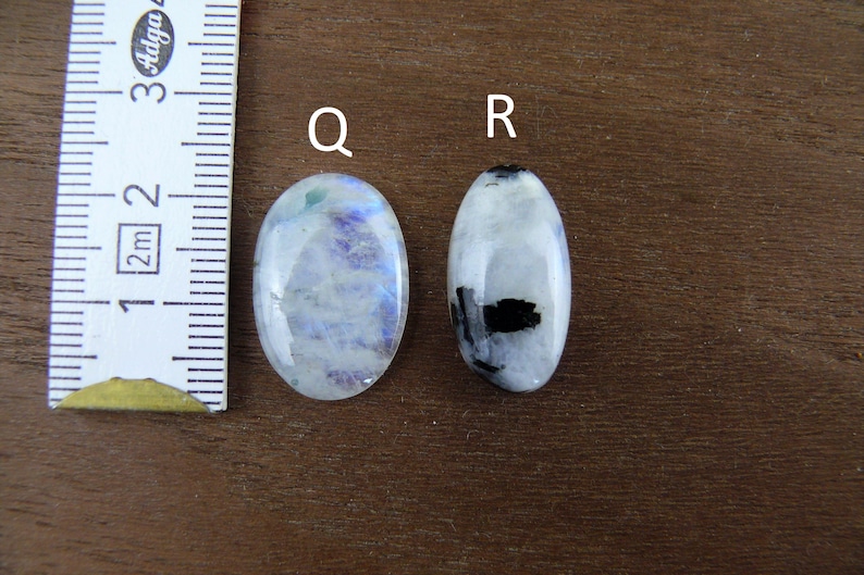 Puede incluir: Dos piedras preciosas de forma ovalada, una con tonos azules y blancos, la otra con inclusiones negras. Se coloca una regla junto a las piedras para comparar el tama&ntilde;o. Las piedras preciosas est&aacute;n sobre una superficie marr&oacute;n.