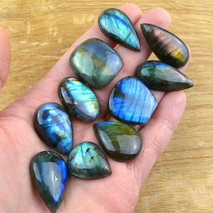Puede incluir: Una colección de piedras preciosas de labradorita pulidas en varias formas, incluyendo lágrimas y óvalos. Las piedras muestran destellos iridiscentes de azul, verde y dorado, con una superficie lisa y reflectante. Las piedras preciosas se sostienen en una mano.