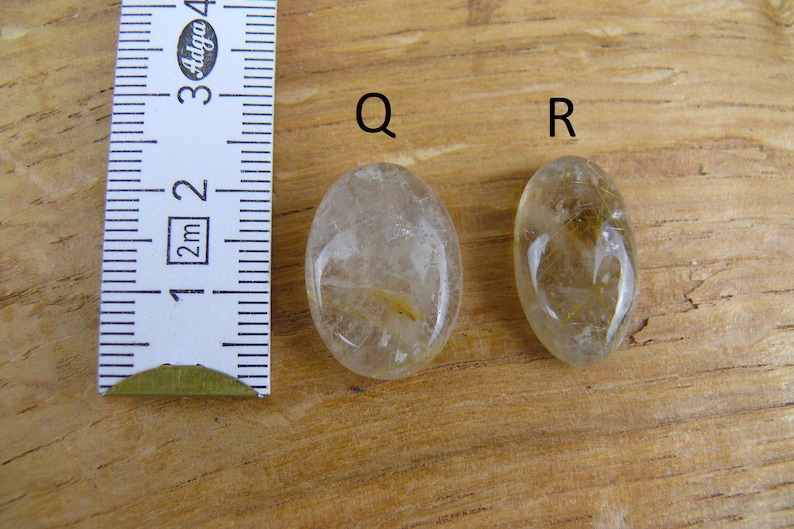 May include: Two oval-shaped clear quartz stones with gold inclusions. The stones are labeled "Q" and "R". A ruler is in the background.