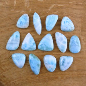 Könnte beinhalten: Eine Sammlung polierter Larimar-Edelsteine in verschiedenen Formen, darunter Tropfen, Ovale und Dreiecke. Die Steine zeigen eine Reihe von Blautönen mit weißer Marmorierung, auf einer warmen Holzoberfläche.