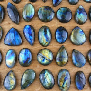 May include: A collection of polished labradorite gemstones in various shapes including ovals, teardrops, and triangles. Each stone displays a dark gray base with iridescent flashes of blue, gold, and green light reflecting from the surface.