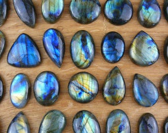 May include: A collection of polished labradorite gemstones in various shapes including ovals, teardrops, and triangles. Each stone displays a dark gray base with iridescent flashes of blue, gold, and green light reflecting from the surface.