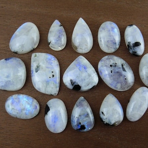 Puede incluir: Una colecci&oacute;n de piedras preciosas de piedra lunar pulidas en varias formas, incluyendo ovaladas, de l&aacute;grima e irregulares. Las piedras muestran una base blanca lechosa con inclusiones azules y negras, sobre una superficie de madera marr&oacute;n.