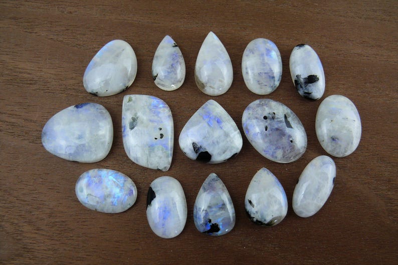 Puede incluir: Una colecci&oacute;n de piedras preciosas de piedra lunar pulidas en varias formas, incluyendo ovaladas, de l&aacute;grima e irregulares. Las piedras muestran una base blanca lechosa con inclusiones azules y negras, sobre una superficie de madera marr&oacute;n.