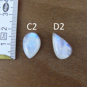 May include: Two pear-shaped moonstone cabochons labeled C2 and D2, positioned next to a white measuring tape showing markings in centimeters. The stones are translucent white with blue adularescence and smooth, polished surfaces.