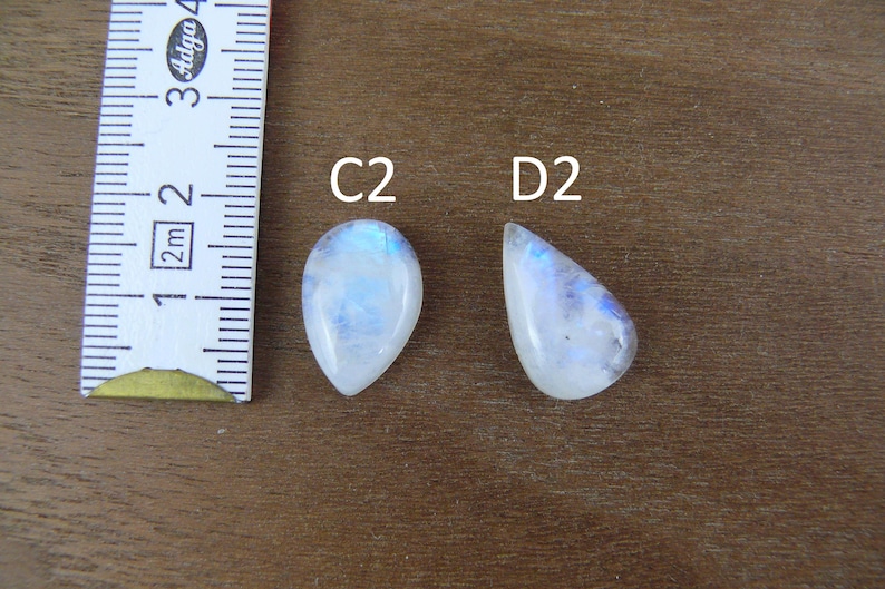 May include: Two pear-shaped moonstone cabochons labeled C2 and D2, positioned next to a white measuring tape showing markings in centimeters. The stones are translucent white with blue adularescence and smooth, polished surfaces.