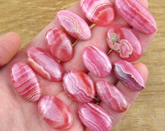 May include: A collection of twelve polished, oval-shaped rhodochrosite gemstones. Each stone features a distinct pattern of vibrant pink and white bands, with smooth, glossy surfaces that highlight the natural mineral layers and swirls.