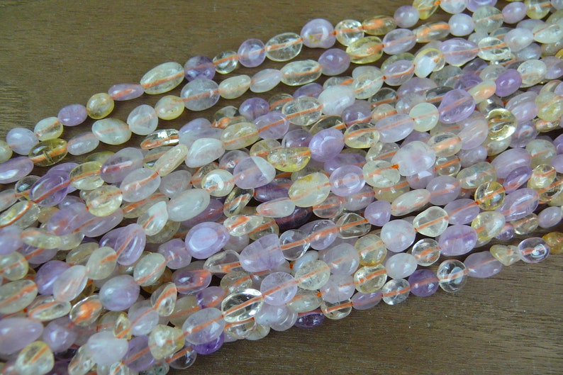 May include: A strand of mixed gemstone beads, featuring a mix of light purple amethyst, clear quartz, and yellow citrine beads. The beads are smooth and oval-shaped, and they are strung on a clear elastic cord.