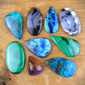 Può includere: Una collezione di nove pietre preziose levigate di varie forme e dimensioni, tra cui ovali, a goccia e forme irregolari. Le pietre presentano colori e motivi diversi, come blu vibrante, verde intenso, viola e bianco traslucido con inclusioni nere.