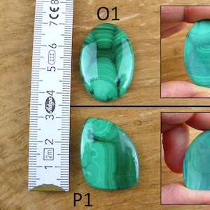 May include: Two polished malachite gemstones, one oval and one irregularly shaped, displayed on a wooden surface. The oval stone is approximately 2 inches long. The malachite has a vibrant green color with swirling patterns. A ruler is placed next to the stones.
