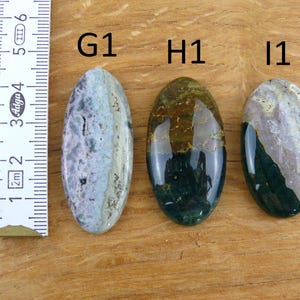 Op de afbeelding: Drie ovaalvormige gepolijste edelstenen met de labels G1, H1 en I1, geplaatst naast een liniaal met centimeters. De stenen hebben gevarieerde patronen in paarse, groene, bruine en cr&egrave;mekleurige tinten met een gladde, glanzende afwerking.
