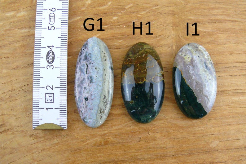 Op de afbeelding: Drie ovaalvormige gepolijste edelstenen met de labels G1, H1 en I1, geplaatst naast een liniaal met centimeters. De stenen hebben gevarieerde patronen in paarse, groene, bruine en cr&egrave;mekleurige tinten met een gladde, glanzende afwerking.