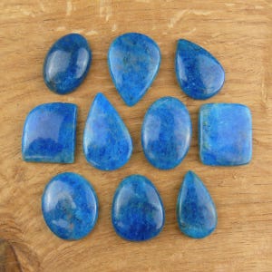 Könnte beinhalten: Eine Sammlung von zehn polierten blauen Edelsteinen in verschiedenen Formen, darunter oval, tropfenförmig und quadratisch. Die Edelsteine haben eine durchscheinende Qualität und liegen auf einer hellbraunen Holzoberfläche.