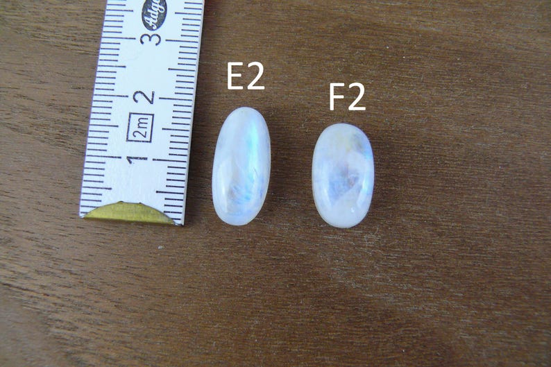 May include: Two oval-shaped, translucent white moonstones labeled E2 and F2, positioned next to a white measuring tape showing metric units. The stones exhibit a subtle blue adularescence and smooth, polished surfaces for jewelry making.