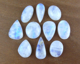 May include: A collection of ten polished moonstone cabochons in various shapes including oval, round, and teardrop. Each gemstone displays a translucent white appearance with iridescent blue flashes visible across the smooth, domed surfaces.