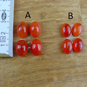 Op de afbeelding: Close-up van twee sets ovaalvormige, gepolijste, oranjerode edelstenen. Een liniaal met centimeter- en millimeter-markeringen staat links. De edelstenen zijn gemarkeerd met A en B. De edelstenen zijn ongeveer 2,5 cm lang.