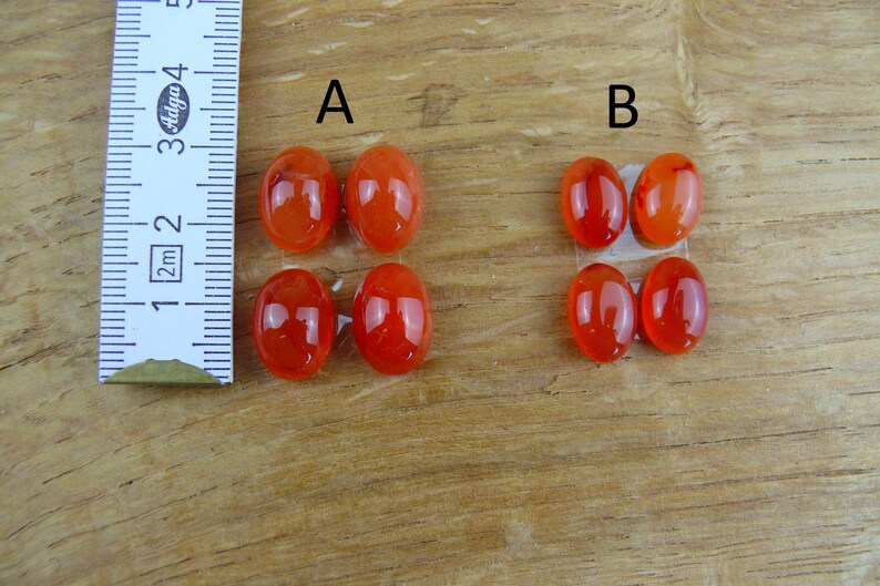 Op de afbeelding: Close-up van twee sets ovaalvormige, gepolijste, oranjerode edelstenen. Een liniaal met centimeter- en millimeter-markeringen staat links. De edelstenen zijn gemarkeerd met A en B. De edelstenen zijn ongeveer 2,5 cm lang.