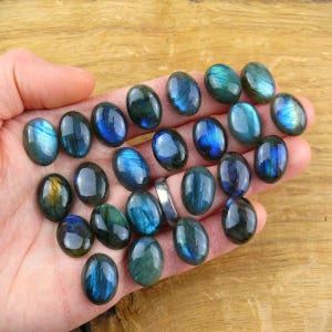 Pode incluir: Uma coleção de pedras preciosas de labradorita ovais em várias tonalidades de azul, verde e dourado. As pedras polidas são seguradas em uma mão, mostrando suas qualidades iridescentes. As pedras preciosas são exibidas em uma superfície de madeira.