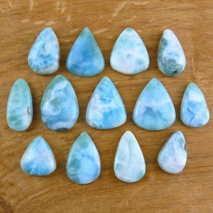 Könnte beinhalten: Eine Sammlung tropfen- und dreieckförmiger Larimar-Edelsteine in Blau- und Weißtönen, angeordnet auf einer Holzoberfläche. Die Steine variieren in der Größe und weisen einzigartige Muster auf.