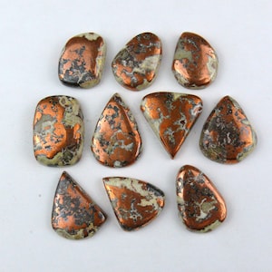 Può includere: Dieci cabochon lucidati caratterizzati da una superficie color rame metallizzato con inclusioni di pietra grigia e bianca screziata. Le pietre presentano varie forme, tra cui gocce, triangoli e rettangoli arrotondati con superfici lisce e riflettenti.