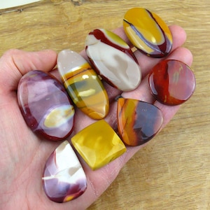 Può includere: Una collezione di pietre lucide di varie forme e dimensioni, tenute in una mano. Le pietre mostrano una gamma di colori, tra cui bordeaux, giallo, crema e marrone. Le pietre sono di forma ovale, a goccia e quadrata.