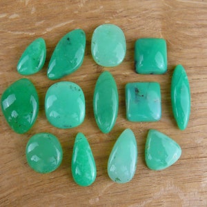 Op de afbeelding: Een verzameling van 12 groene edelsteen cabochons in verschillende vormen en maten. De cabochons zijn gerangschikt op een houten oppervlak.