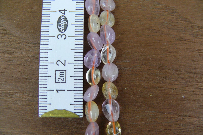 May include: Two strands of beads, one strand is light pink and the other is light yellow. The beads are oval shaped and are strung on a thin orange thread. A ruler is in the background to show the size of the beads.