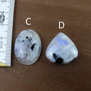 Puede incluir: Dos cabujones pulidos de piedra lunar, uno ovalado y otro en forma de l&aacute;grima, con inclusiones negras y un brillo nacarado. Una regla se coloca junto a las piedras, mostrando medidas en cent&iacute;metros. Las piedras est&aacute;n sobre una superficie de madera marr&oacute;n.