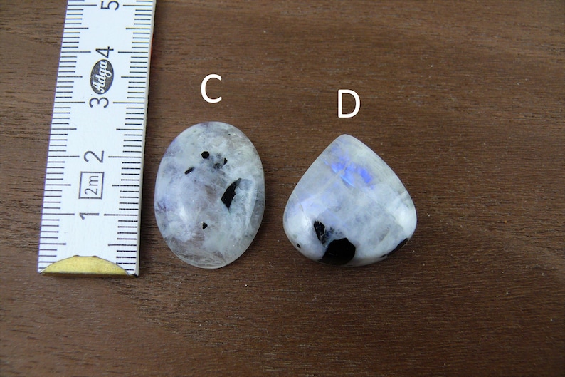 Puede incluir: Dos cabujones pulidos de piedra lunar, uno ovalado y otro en forma de l&aacute;grima, con inclusiones negras y un brillo nacarado. Una regla se coloca junto a las piedras, mostrando medidas en cent&iacute;metros. Las piedras est&aacute;n sobre una superficie de madera marr&oacute;n.