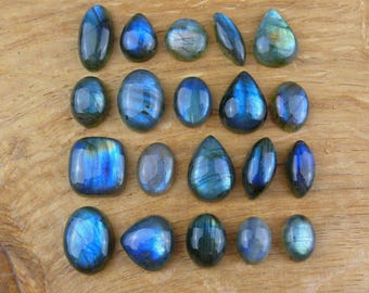 Puede incluir: Una colección de piedras preciosas de labradorita pulidas en varias formas, incluyendo ovaladas, en forma de lágrima y cuadradas. Las piedras muestran destellos iridiscentes azules y verdes sobre un fondo oscuro, sobre una superficie de madera.
